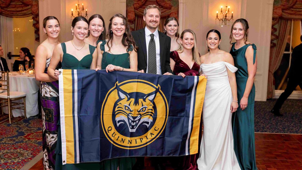 Quinnipiac alumni pose for a picture holding a Quinnipiac University banner at a wedding