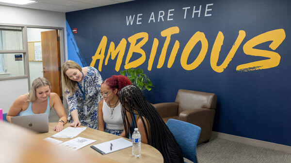 Three students and a professor review a resume with a wall behind them saying we are the ambitious