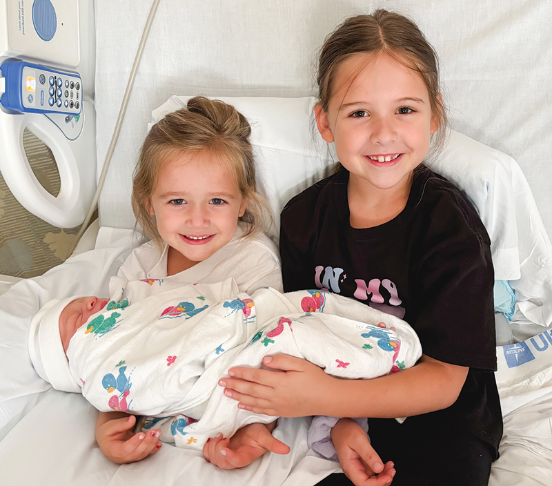 Two little girls hold their new-born brother on a hospital bed